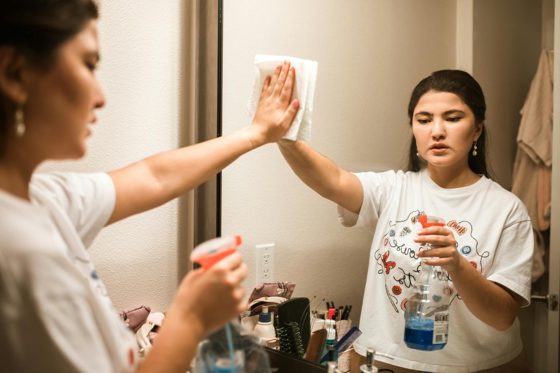 Woman wiping a bathroom mirror with a paper towel while holding a spray bottle