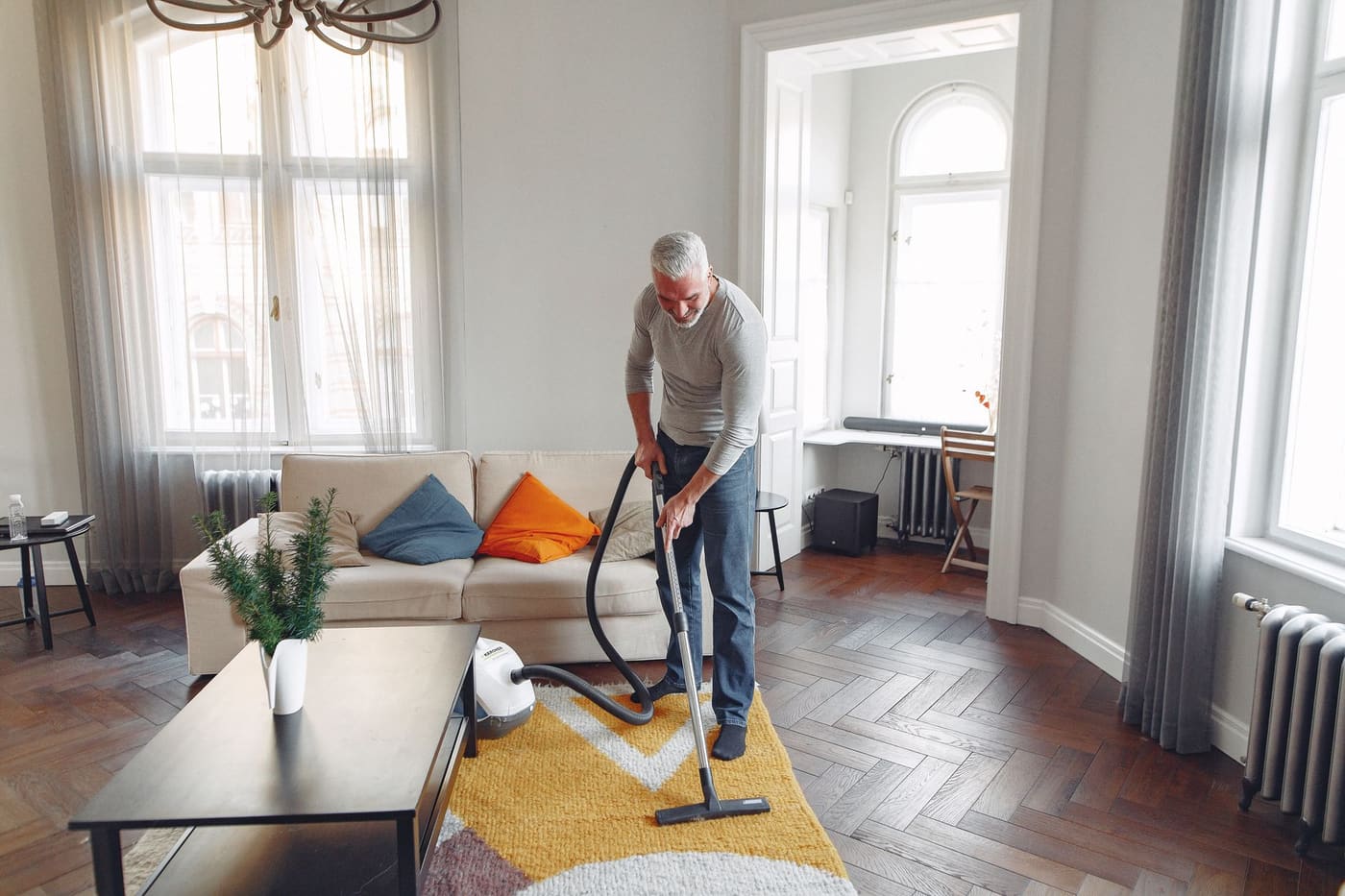 Man vacuuming a rug in a bright living room with a sofa and tall windows