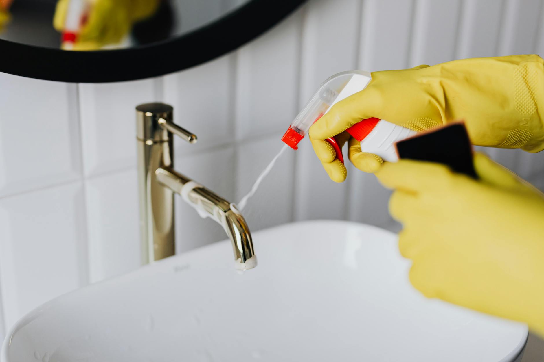 Gloved hand spraying cleaner onto a chrome bathroom faucet above a white sink